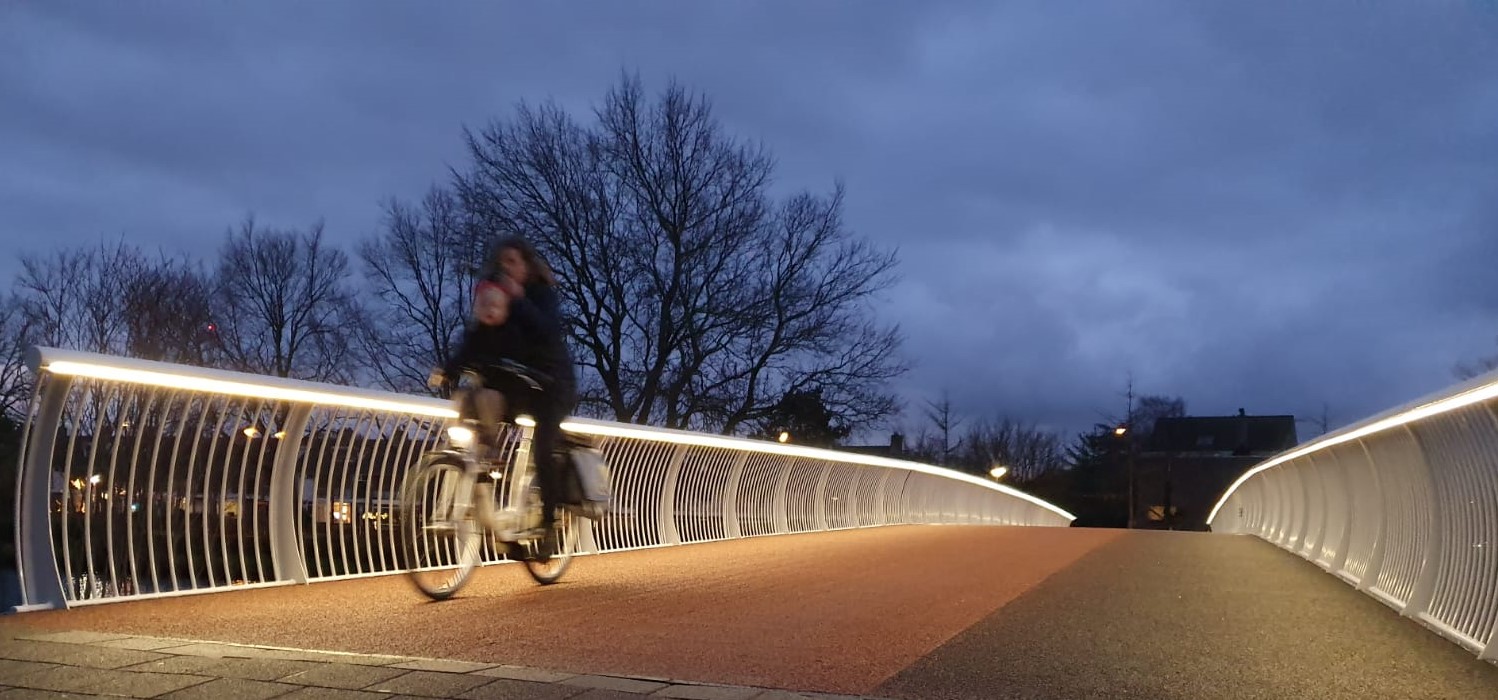 Moeder met kind fietst veilig verlicht over de Jan Hendrik Oort brug in Oegstgeest. De brugverlichting wordt gerealiseerd door de de verlichte brugleuning. Het maatwerk stalen ILLUNOX armatuur is op locatie door vakmensen voorzien van de ledverlichting. Een veilige brug door een functionele verlichting met asymmetrische lichtoutput op het te belopen oppervlak van de voetgangers- en fietsersbrug.